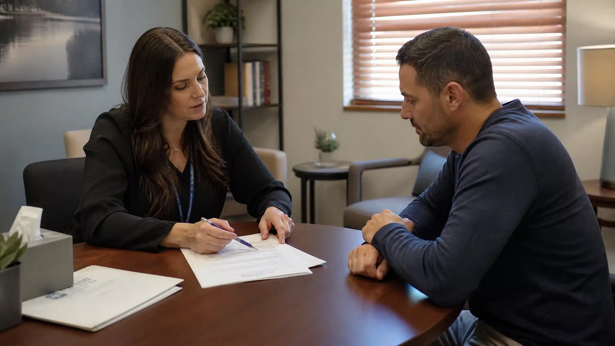 Adult meeting with an admissions counselor at a treatment center, reviewing intake paperwork together, calm expressions, realistic professional setting.